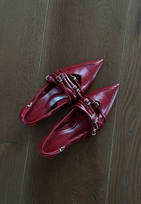 Κόκκινα δερμάτινα παπούτσια με μυτερές μύτες διαθέτουν σχέδιο σταυρωτών λουριών με ασημένιες αγκρέδες. Η λεία υφή και το ανοιχτό πίσω σχέδιο ενισχύουν την αναπνοή.