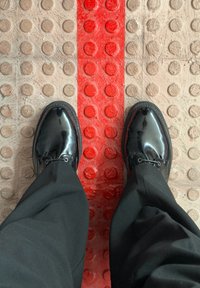 Des chaussures noires vernies et un pantalon se tiennent sur un trottoir texturé avec une bande de guidage tactile rouge pour les piétons malvoyants.