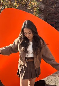 Cardigan marron tricoté avec des manches surdimensionnées porté sur un top blanc, associé à une jupe à carreaux. Fond abstrait orange vif.