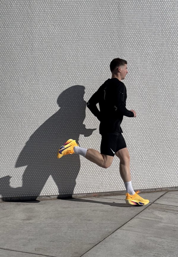 Mann iført svarte treningsklær og knallgule joggesko løper forbi en teksturert hvit vegg som kaster en tydelig skygge.