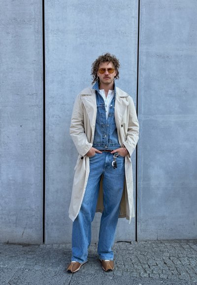 Hombre con pelo rizado y gafas de sol que lleva un abrigo trench beige, chaqueta vaquera, vaqueros azules y zapatos marrones, de pie contra una pared de hormigón gris.