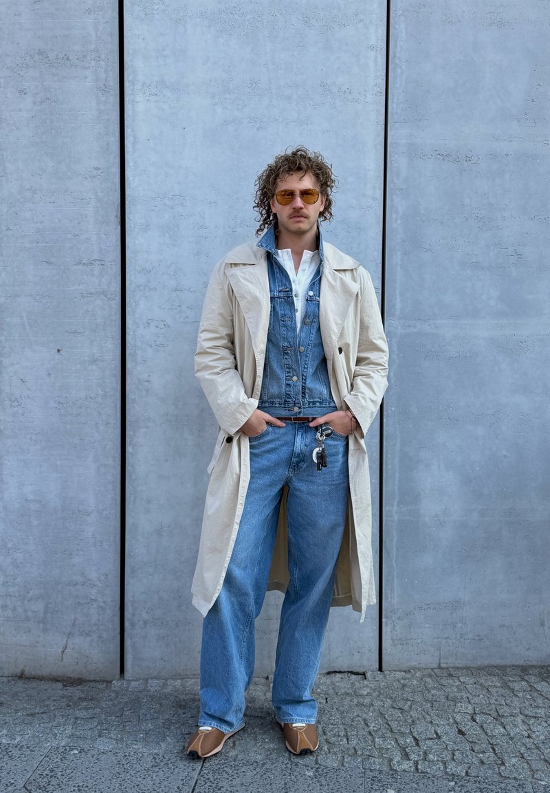 Man met krullend haar en zonnebril, gekleed in een beige trenchcoat, spijkerjasje, blauwe jeans en beige schoenen, staand tegen een grijze betonnen muur.