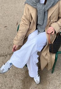 Beige Trenchcoat über einem grauen Pullover, weißen Hosen und weißen Sneakers mit schwarzen Streifen. Eine gemusterte Handtasche ruht auf einem grünen Stuhl.