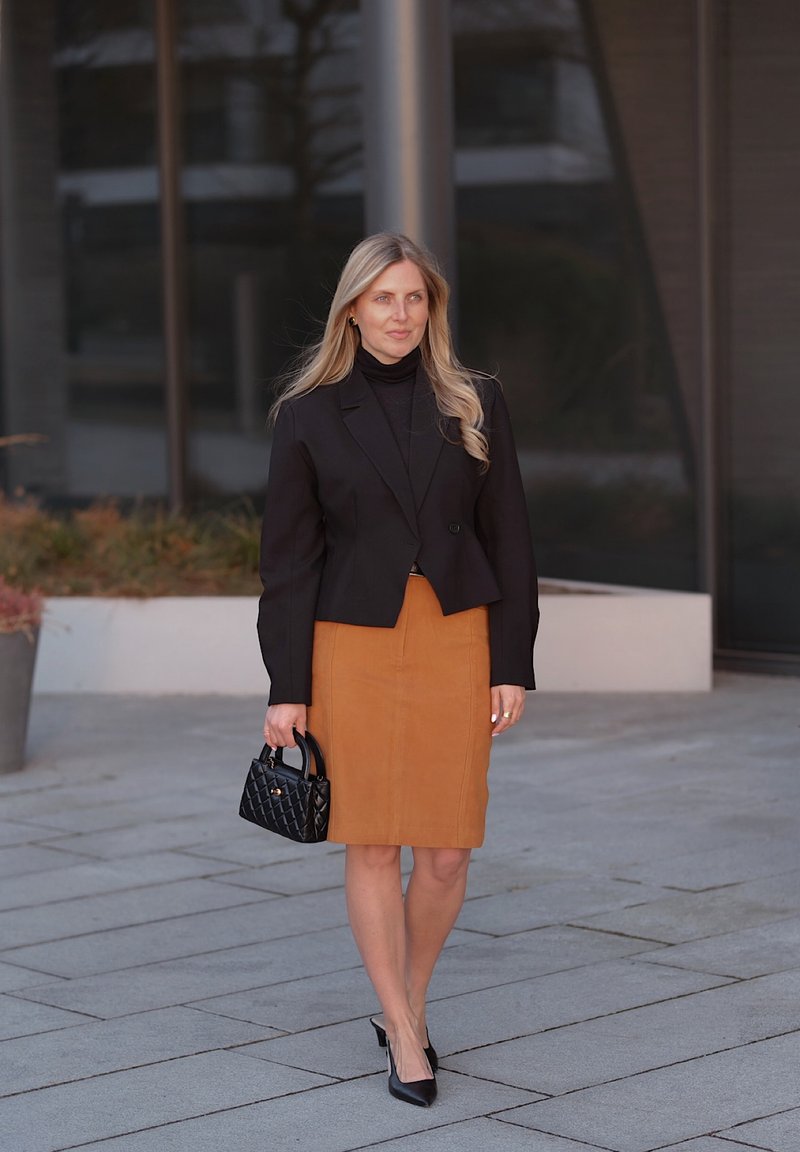 Femme blonde marchant en plein air portant un blazer noir, un top noir, une jupe beige longueur genou, des talons noirs, et portant un petit sac à main noir.