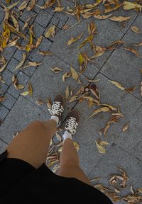 Pantofi sport maro cu șireturi albe și tălpi gri, purtați cu șosete albe, deasupra unui paviment gri presărat cu frunze uscate galbene și maro.