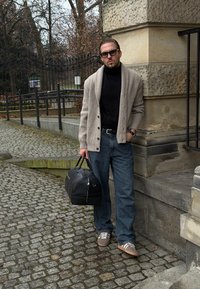 Cardigan côtelé beige clair par-dessus un col roulé noir, jean bleu et baskets grises. Tenant un sac de sport noir, debout sur une rue pavée.