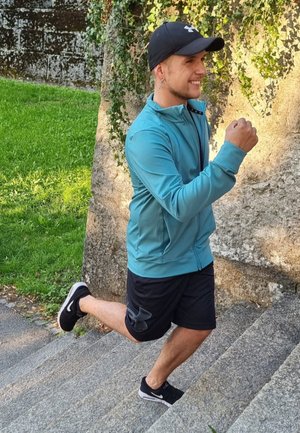 Hombre corriendo por escalones de piedra, vestido con una chaqueta de cremallera en color verde azulado y pantalones cortos negros. Zapatillas deportivas negras con detalles blancos, sonriente, con vegetación de fondo.