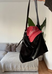 Μαύρη δερμάτινη τσάντα ώμου που κρατιέται με το ένα χέρι, περιέχει ένα κόκκινο καπέλο μπέιζμπολ με λευκό κείμεν ο, μέσα σε ένα σαλόνι με καναπέ και φυτό.