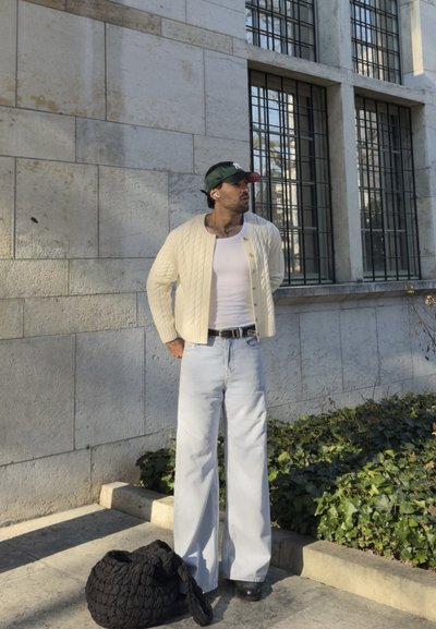 Hombre con cárdigan crema, camiseta blanca sin mangas, vaqueros claros y gorra verde, de pie junto a una pared de piedra y una ventana, bolsa tejida negra en el suelo.