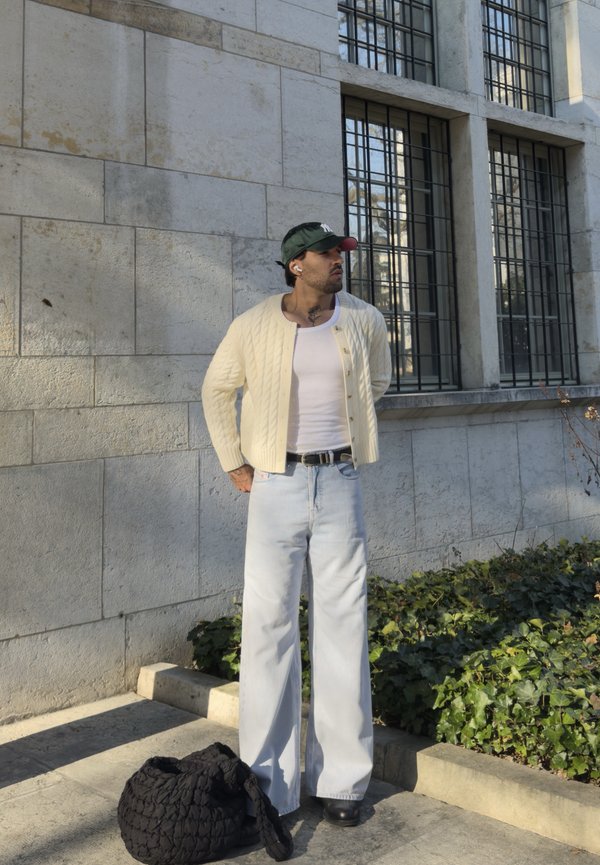 Mann i kremfarget cardigan, hvit singlet, lyse jeans og grønn caps står ved en steinvegg og vindu, svart vevd veske på bakken.