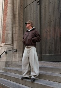 Un homme portant un pantalon beige surdimensionné, une veste marron et une casquette à motif se tient sur des marches en pierre près d'une grande porte en bois et d'un mur de briques.