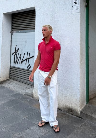 Camisa polo roja de manga corta, pantalones holgados blancos, chanclas negras, de pie frente a una pared blanca con grafitis y una persiana metálica detrás.