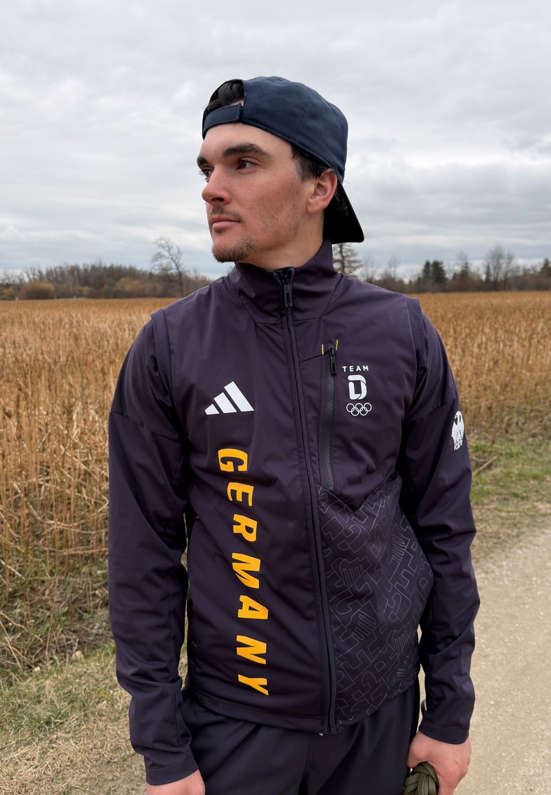 Jeune homme portant une veste olympique allemande bleu marine et une casquette à l'envers, debout sur un sentier à côté d'un champ sous un ciel nuageux.
