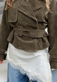 Veste ceinturée en daim vert olive portée sur une écharpe blanche bordée de dentelle et un jean bleu, sur une personne aux cheveux bouclés dont des sections sont visibles.