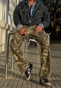 Chaqueta de mezclilla sobre una camisa a rayas, combinada con pantalones cargo de camuflaje holgados y zapatillas negras con detalles blancos. Entorno urbano con barandilla metálica.