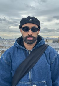 Zils džinsi jakas ar pūkainu apkakli, melns bere un melnas saulesbrilles. Cilv�ēkam ir bārda un melna plecu soma pāri torso.