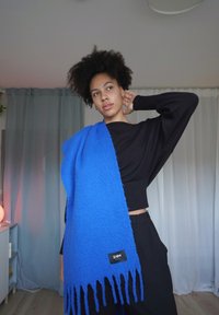 Blå, teksturert skjerf med frynsete kanter, båret over en svart cropped genser. Merke synlig, med merkevarenavn på en svart etikett.