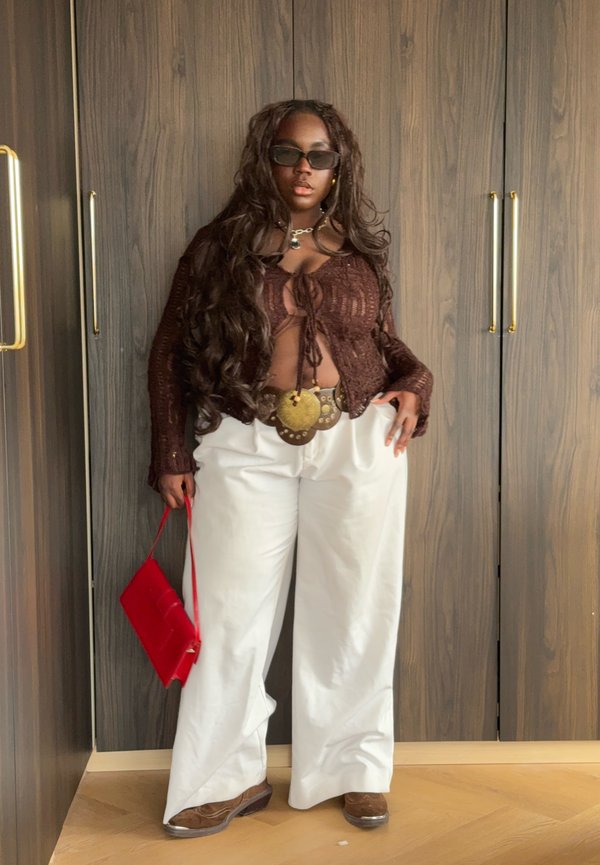 Personne portant un haut en maille marron, un pantalon blanc à jambes larges, une grande ceinture décorative, des lunettes de soleil foncées, tenant un sac à main rouge, debout contre une armoire en bois.