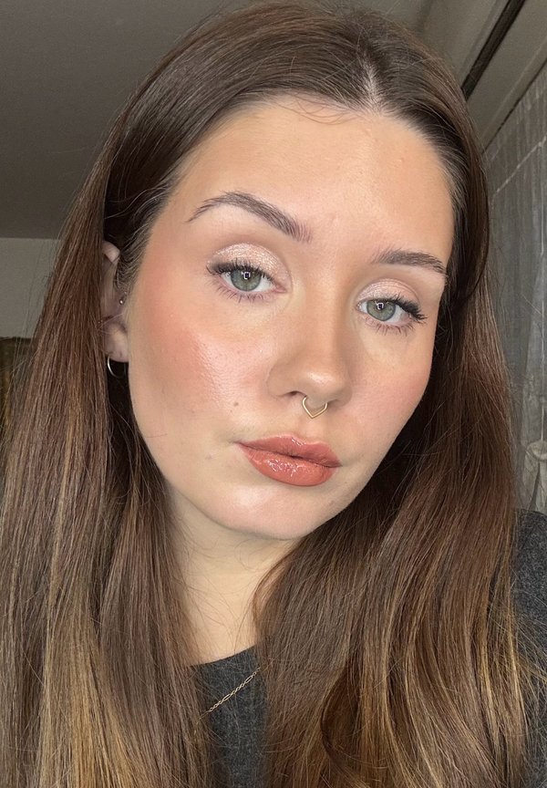 Young woman with long brown hair, glossy peach lipstick, shimmery eyeshadow, subtle blush, and a gold heart-shaped septum ring, wearing a dark top.