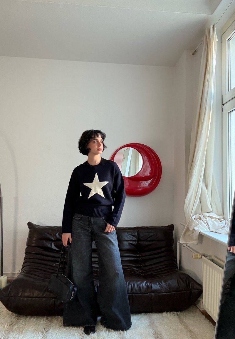 Pull marine avec un motif d'étoile blanche, associé à un jean large foncé. Le mannequin tient un petit sac à main noir, devant un canapé marron.