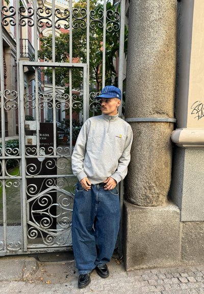 Sudadera gris con cremallera, jeans azules holgados, zapatos negros y una gorra azul. El fondo presenta una reja metálica ornamentada y una pared de piedra.