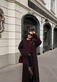 Langer, dunkelbrauner Mantel über einem burgunderroten Pullover, kombiniert mit schwarzen Shorts und Spitzstrumpfhosen. Dazu eine rote Handtasche und Sonnenbrille.