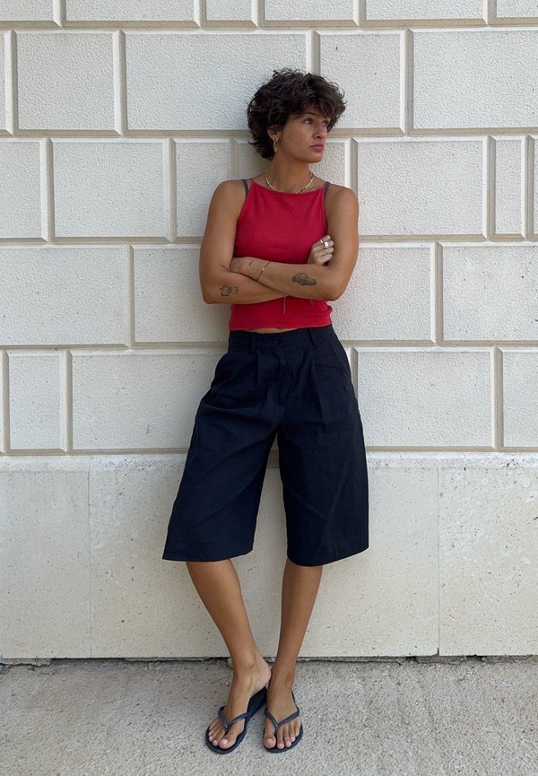 Young person with short curly hair stands with arms crossed wearing a red sleeveless top, black knee-length shorts, and black sandals.