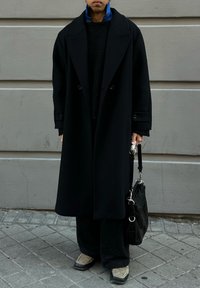 Long black coat with wide lapels, layered over a textured black jumper. Black wide-leg trousers and beige shoes. Black bag with silver details.