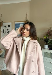 Rosa Wollmischung-Peacoat mit doppelreihigen Knöpfen und breiten Revers, kombiniert mit einem weißen Fellschal. Der Hintergrund zeigt Pflanzen und Kunstwerke.