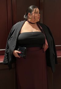 Femme portant un haut noir sans bretelles, une jupe bordeaux, une veste noire posée sur les épaules, tenant une pochette noire, regardant sur le côté.