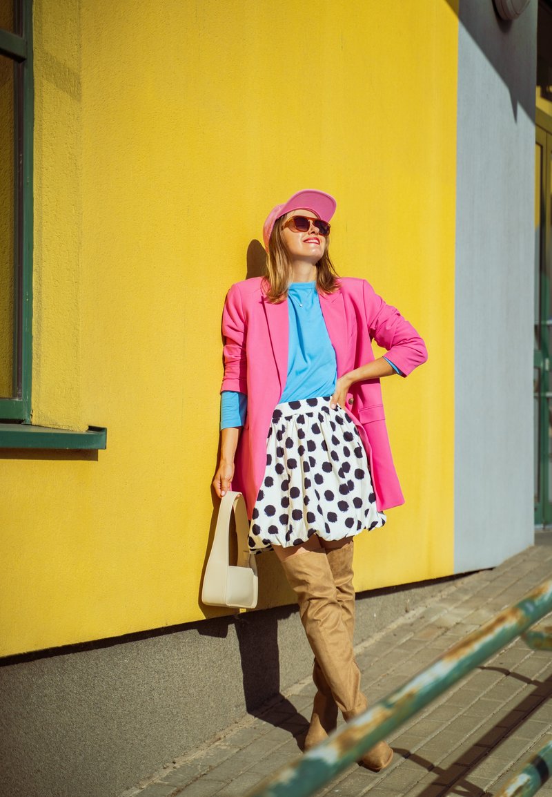 Blazer rosa, blusa azul, saia de bolinhas, botas beijas até ao joelho. A segurar uma mala bege contra uma parede amarela. Óculos de sol e um boné adicionam toques de estilo.