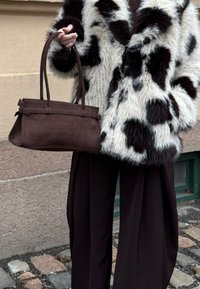 Personne portant une veste en fausse fourrure noire et blanche à pois et un pantalon noir, tenant un grand sac à main en cuir marron à côté d'un mur de pierre.