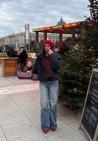 Une femme portant une veste courte grise, un béret rouge et un pull à rayures tient un sac à main. Elle porte un jean large et des chaussures rouges, se tenant en plein air près d'un sapin de Noël.
