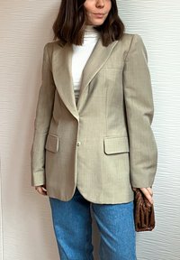 Femme portant un blazer beige sur un col roulé blanc et un jean bleu, tenant une pochette tressée marron devant un mur blanc texturé.
