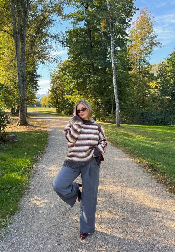 Mujer con suéter de rayas y gafas de sol posando en un camino de grava iluminado por el sol, rodeada de árboles verdes en un parque.