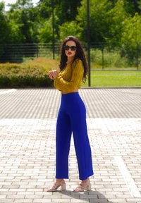 Žlutý texturovaný cropped top s dlouhým rukávem, vysoké modré široké kalhoty a béžové sandály na řemínky na vzorované venkovní dlažbě.