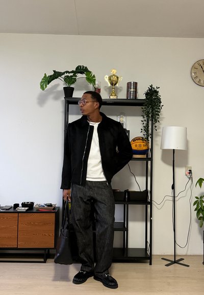 Hombre con chaqueta negra y pantalones grises de pie sosteniendo una bolsa negra en una habitación con estantería negra, balón de baloncesto, plantas y lámpara de pie.