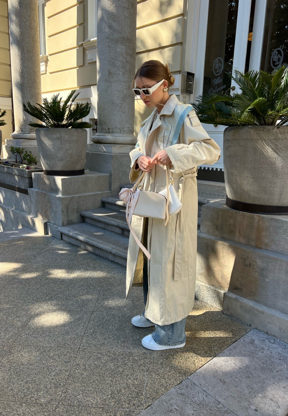 Trenchcoat in un beige chiaro con una cintura, borsa bianca con un fiocco rosa, jeans in denim e sneakers bianche. In piedi accanto a gradini di pietra e vasi.