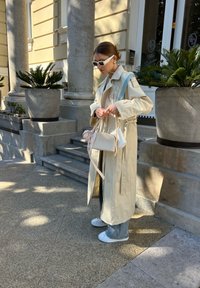 Trenchcoat in un beige chiaro con una cintura, borsa bianca con un fiocco rosa, jeans in denim e sneakers bianche. In piedi accanto a gradini di pietra e vasi.