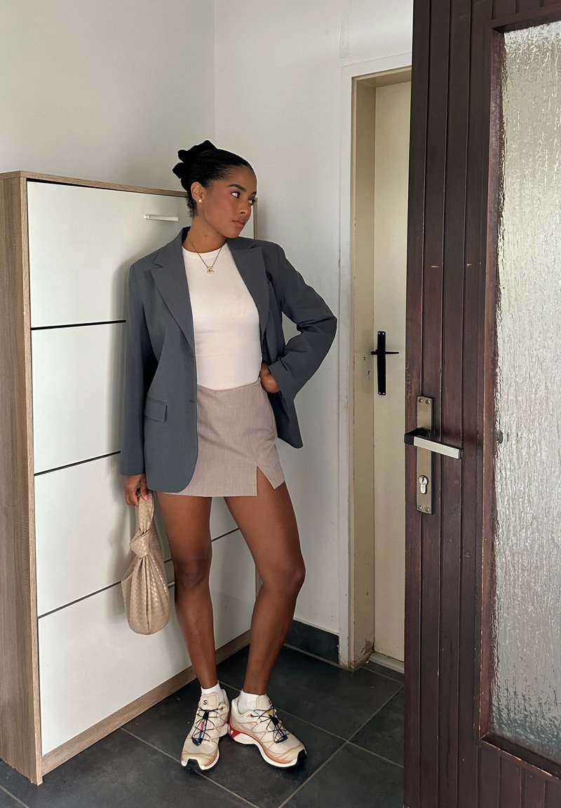 Grey blazer over a white top, teamed with a beige mini skirt. Transparent handbag and multi-coloured trainers. Modern interior setting.