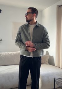 Homme avec barbe et lunettes ajustant sa montre, portant une veste verte, un pull beige et un pantalon noir, debout dans un salon lumineux.