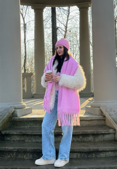 Bufanda de lana rosa con flecos, gorro de punto rosa, abrigo blanco con cuello de piel, vaqueros de mezclilla azul claro y zapatillas blancas frente a escalones de piedra y columnas.