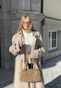 Mladá žena s dlouhými blond vlasy, nosící brýle, béžový trenčkot, bílý pletený svetr, držící hnědousuede kabelku na dlážděné ulici.