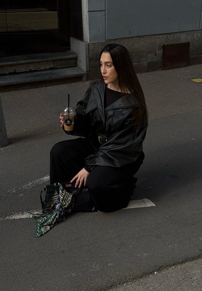 Joven mujer con chaqueta y pantalones de cuero negro arrodillada en la calle, sosteniendo café con hielo, con bolso negro y pañuelo estampado en el suelo.
