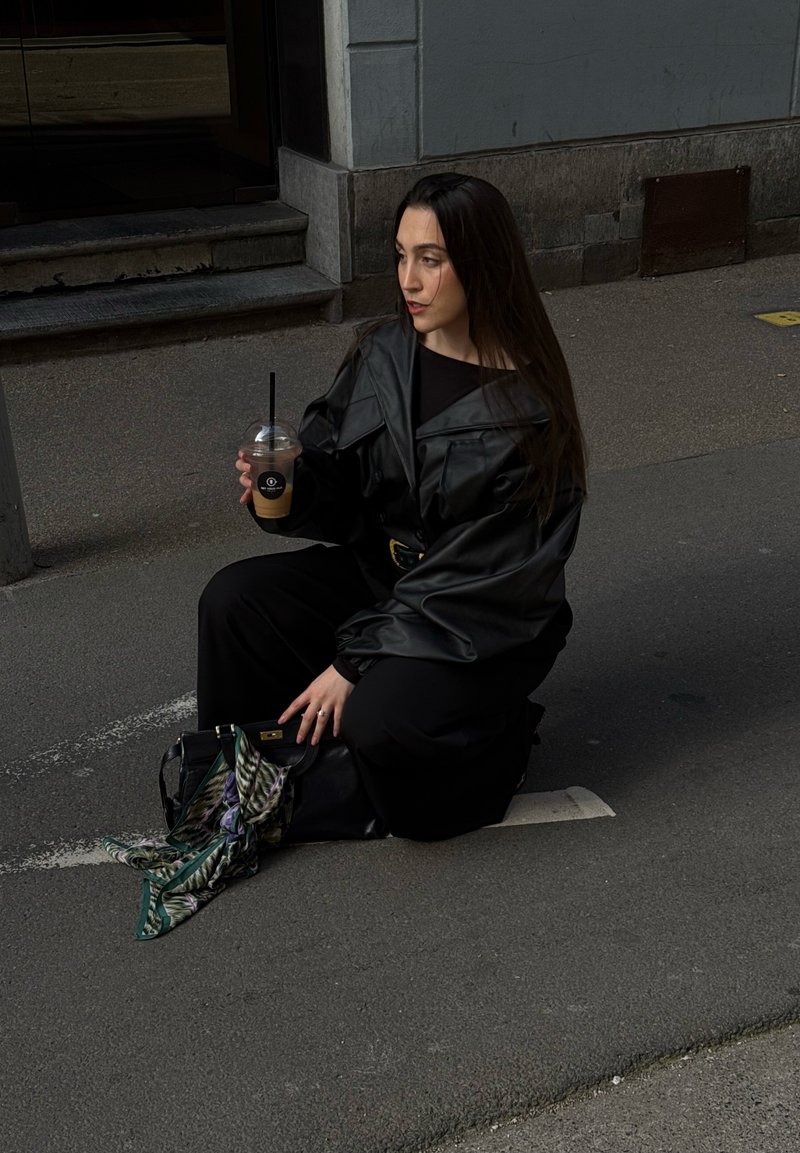 Jeune femme en veste et pantalon en cuir noir à genoux dans la rue, tenant un café glacé, avec un sac à main noir et une écharpe à motifs au sol.