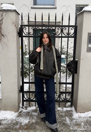 Joven mujer con chaqueta negra, bufanda a rayas, vaqueros oscuros y botas, de pie frente a una puerta de hierro forjado negra con nieve en el suelo.