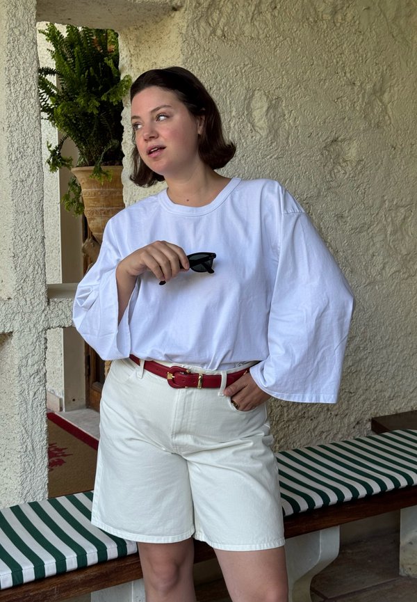 Donna con capelli corti castani che indossa una camicia bianca larga a maniche lunghe, pantaloncini bianchi, cintura rossa, che tiene occhiali da sole neri, in piedi accanto a una panchina a righe.