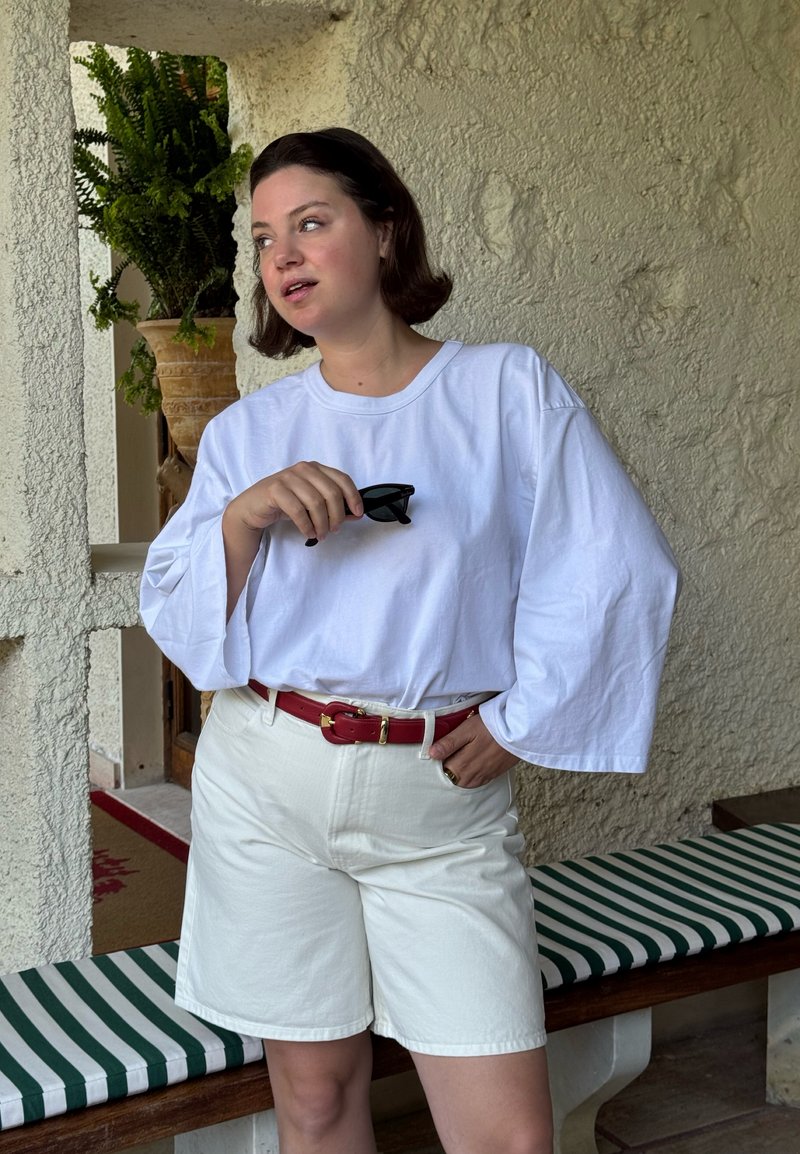 Femme aux cheveux bruns courts portant une chemise blanche ample à manches longues, un short blanc, une ceinture rouge, tenant des lunettes de soleil noires, debout à côté d’un banc rayé.