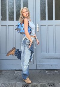 Γκρίζο crop top, μπλε καμουφλάζ τσάντα με καφέ λεπτομέρειες, φθαρμένα ανοιχτό γαλάζια φαρδιά τζιν και καφέ μποτάκια. Στηρίζεται σε μια γκρίζα πόρτα.