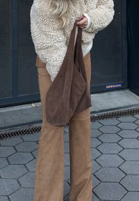 Τσάντα καφέ σουέτ με καρό σχέδιο κρατημένη από ένα άτομο που φοράει μια φουντωτή κρεμ ζακέτα και καφέ σουέτ παντελόνι σε φαρδιά γραμμή, τοποθετημένη σε ένα υφασματοποιημένο μαύρο φόντο.